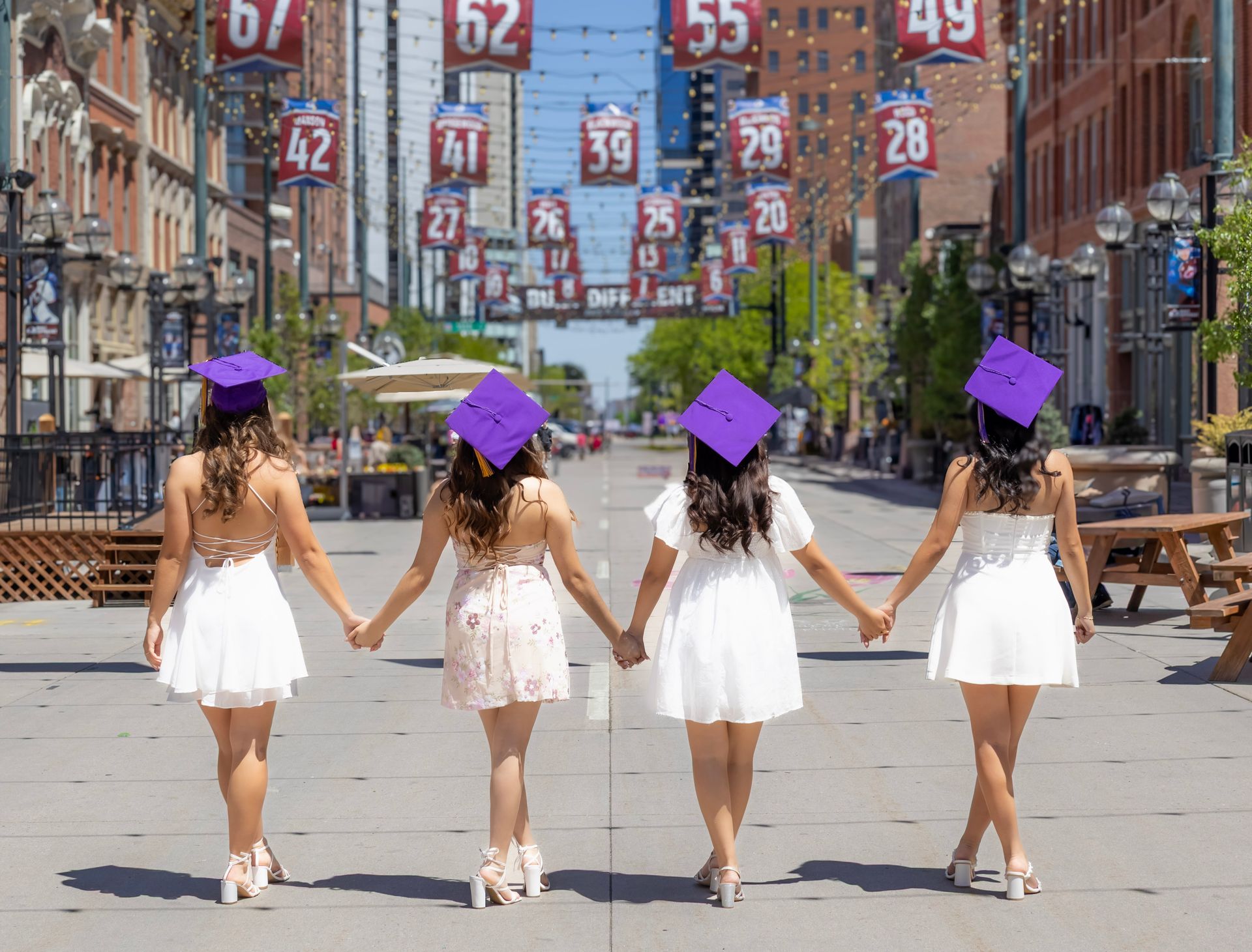 Three female graduates are holding hands while walking down a city street.