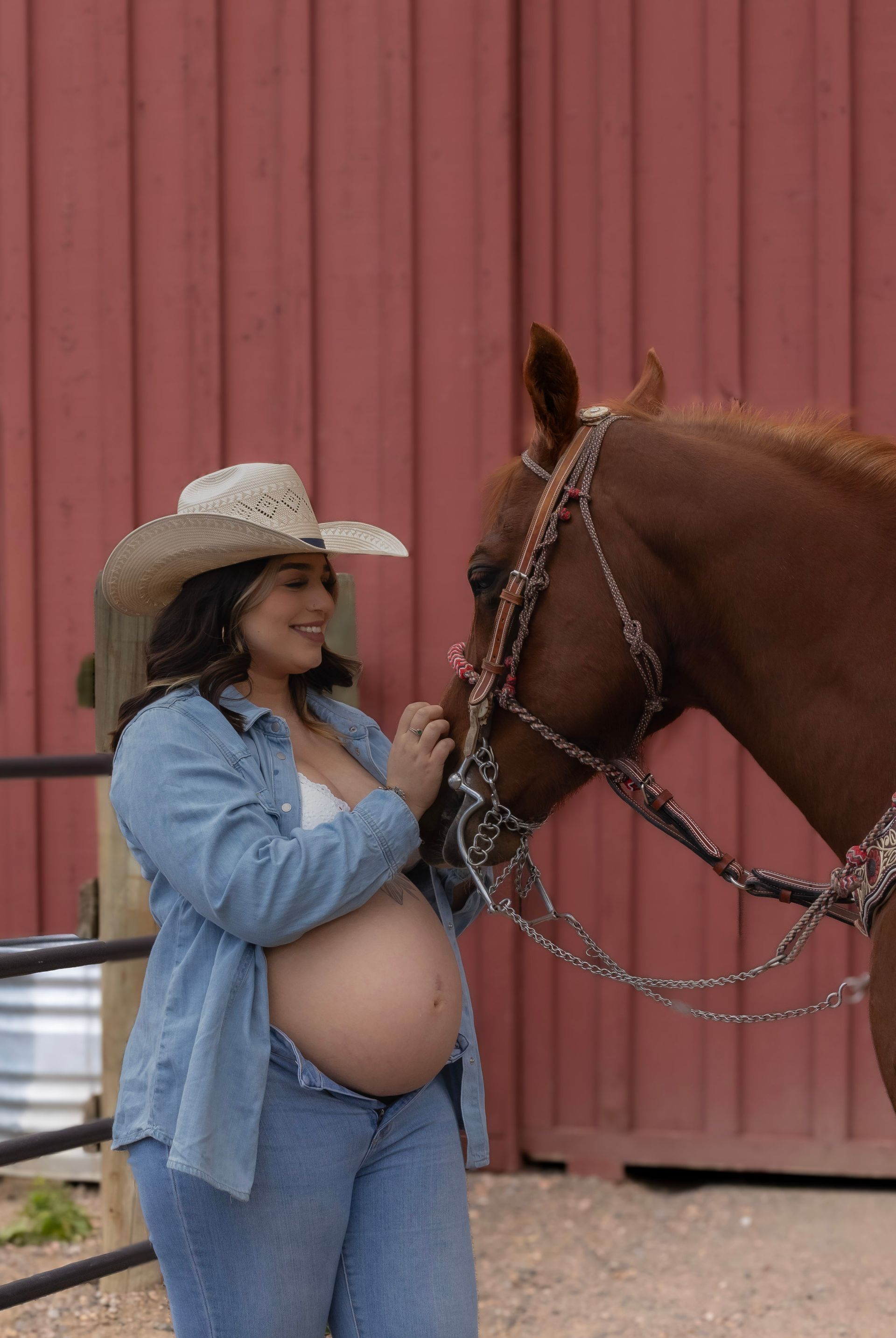 A pregnant woman in a cowboy hat is standing next to a brown horse.