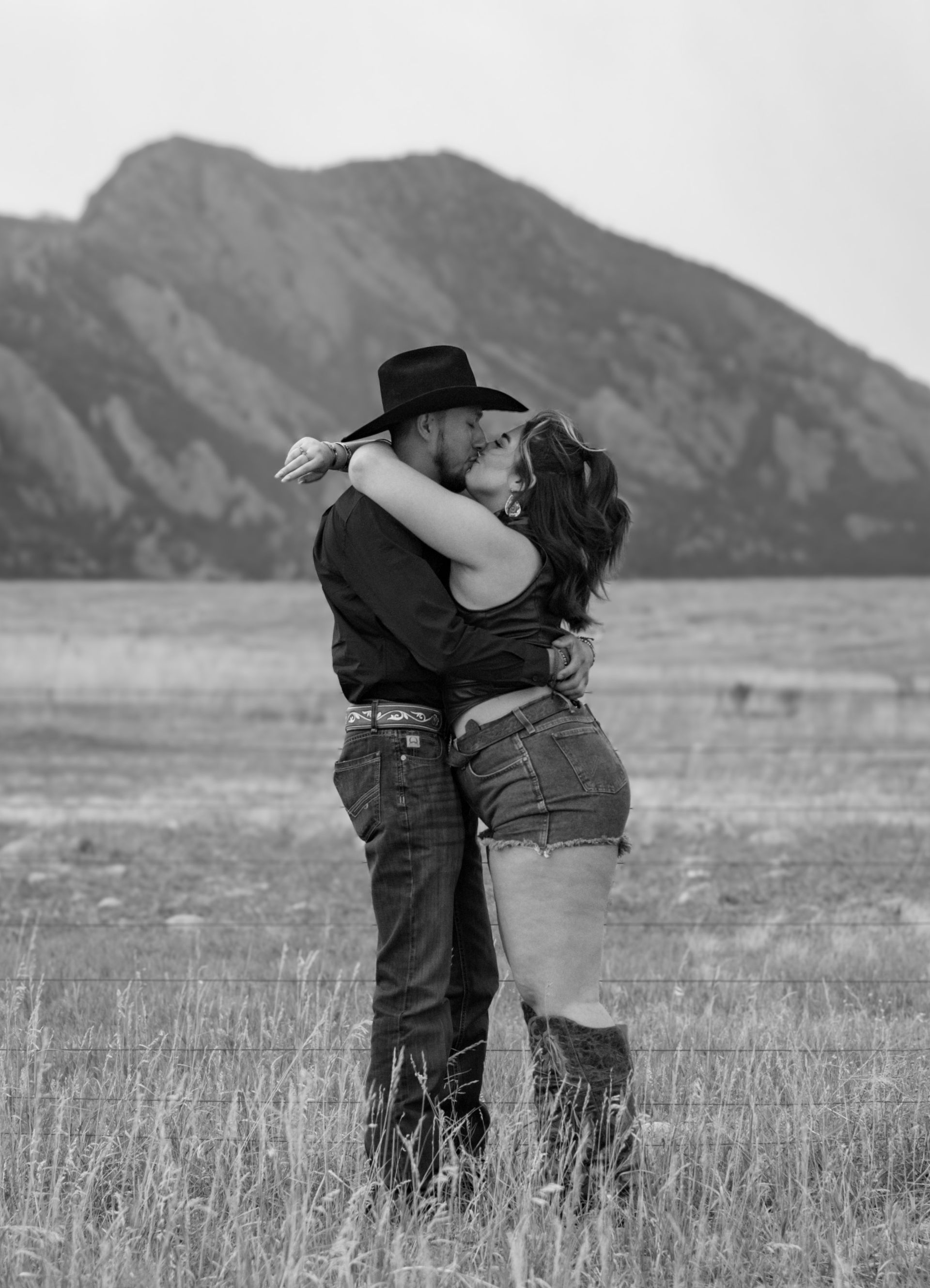 A man and a woman are kissing in a field with mountains in the background.