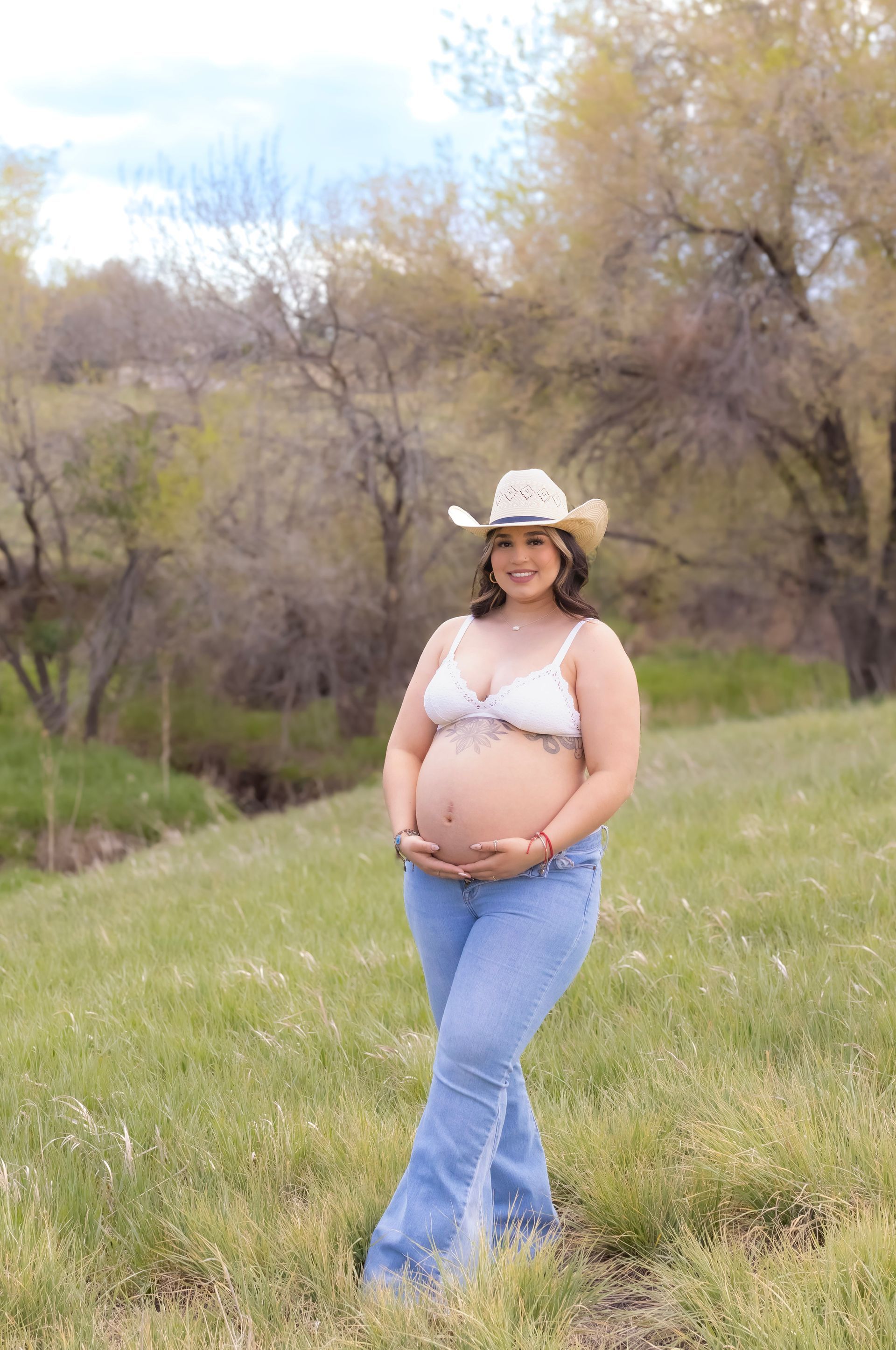 A pregnant woman wearing a cowboy hat and jeans is standing in a field.
