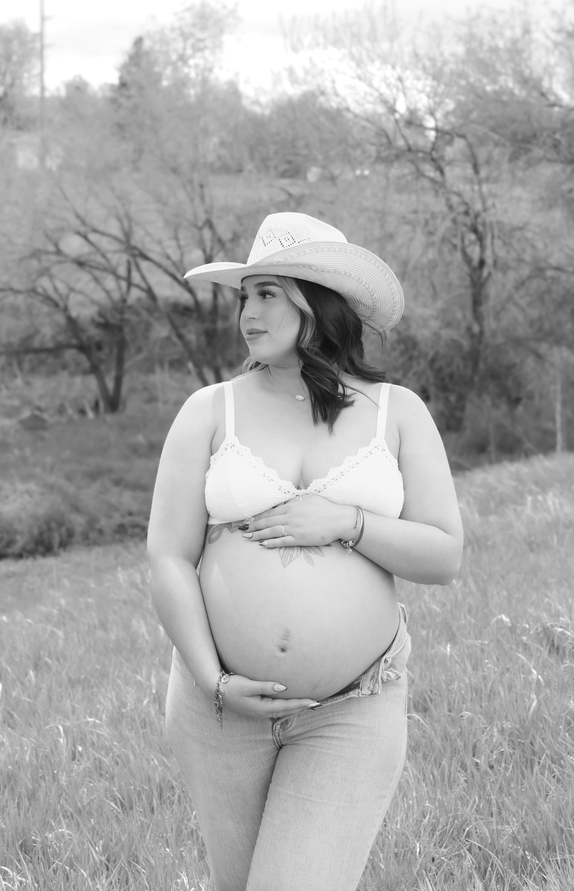 A pregnant woman wearing a cowboy hat is standing in a field holding her belly.