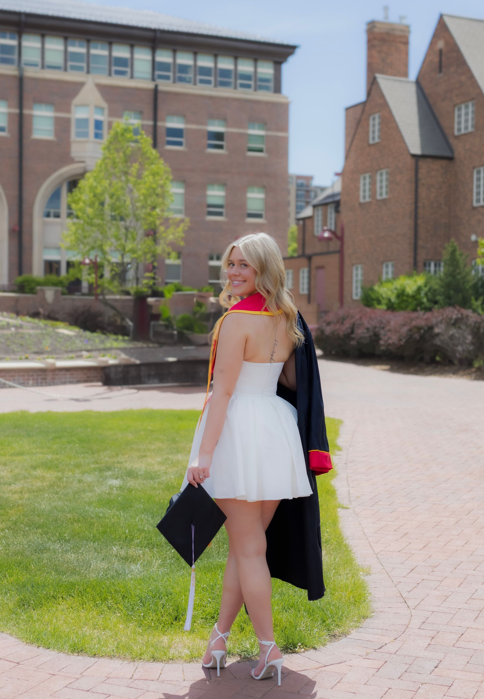 A woman in a graduation cap and gown is standing in front of a brick building.