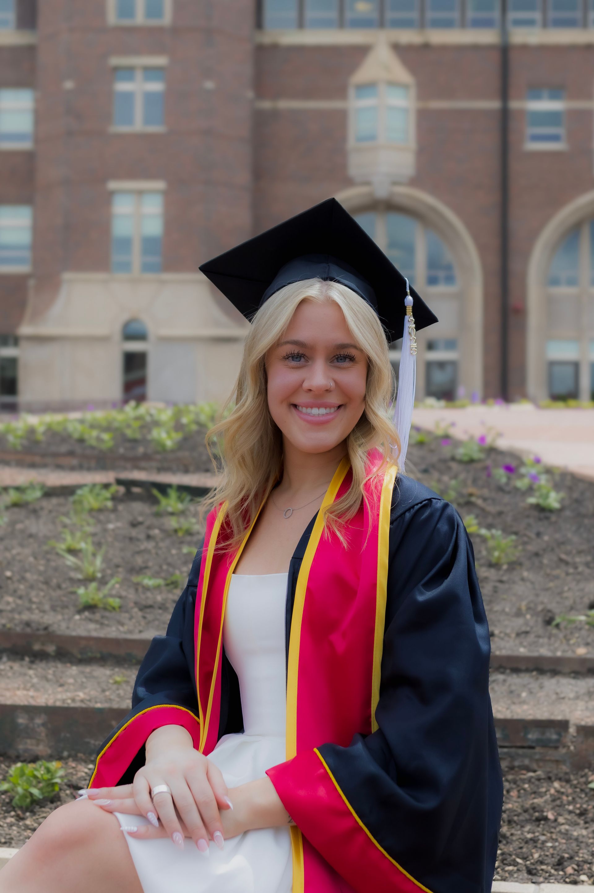 A woman in a graduation cap and gown is sitting in front of a building.
