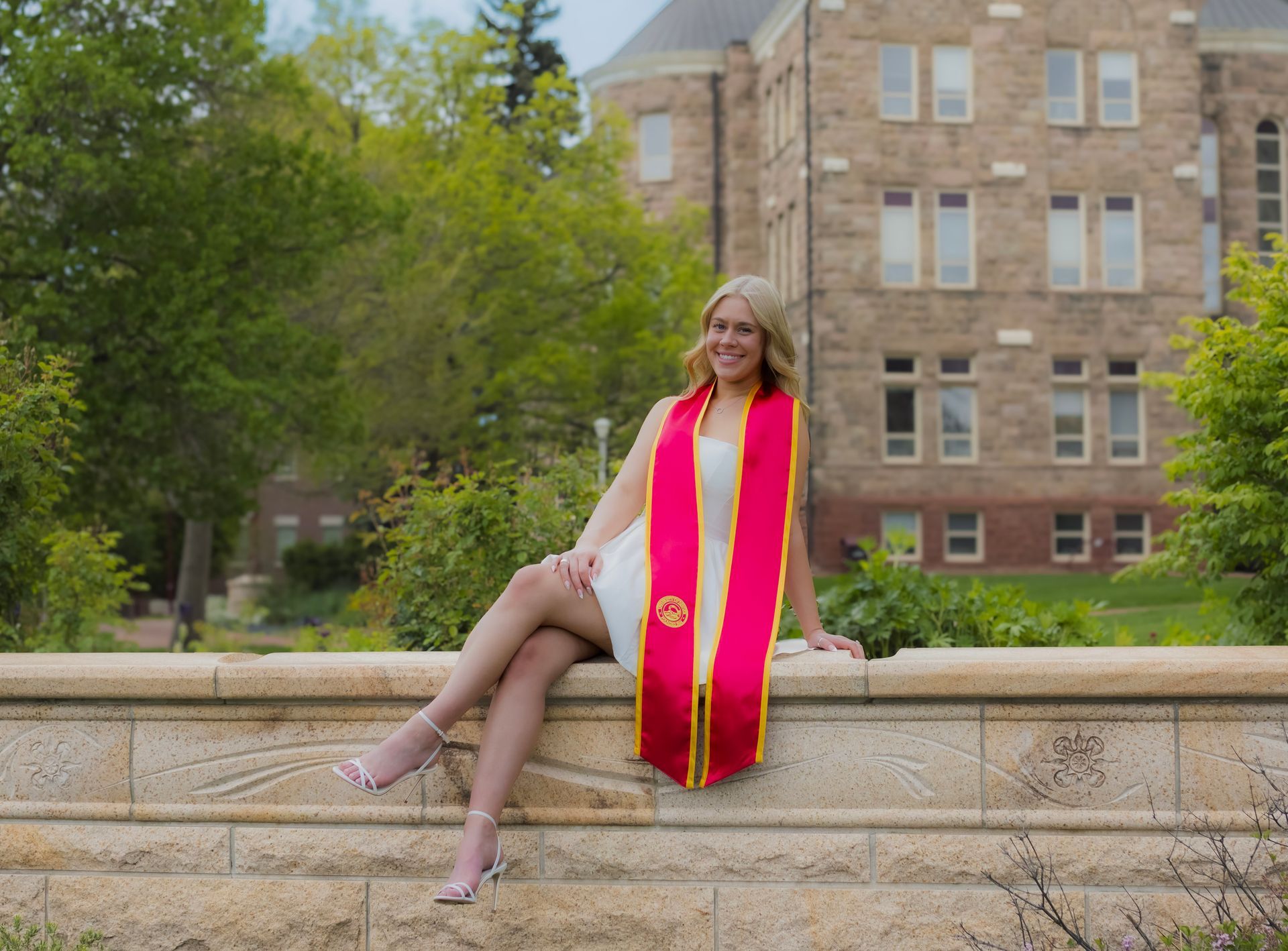 A woman in a graduation cap and gown is sitting on a stone wall.