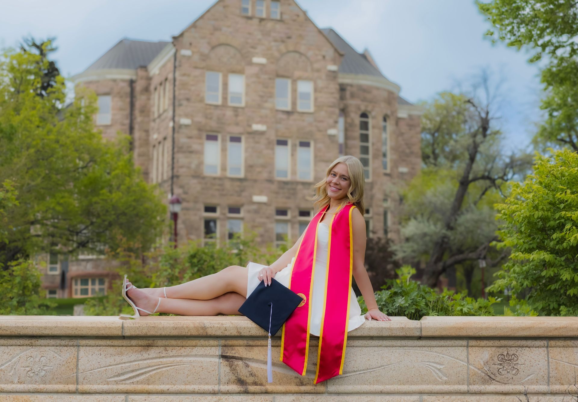 A woman in a graduation cap and gown is sitting on a wall.