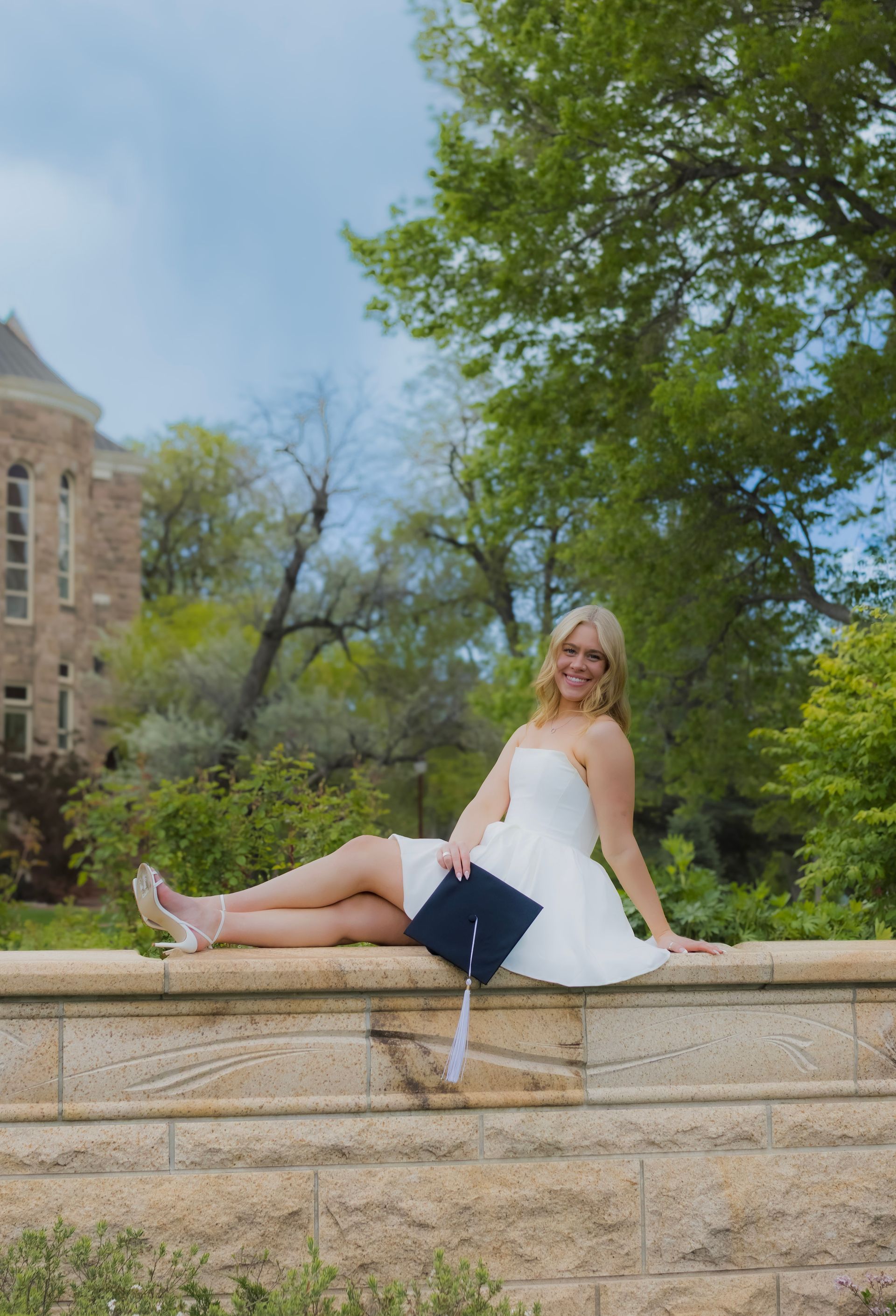 A woman in a white dress is sitting on a stone wall with her legs crossed.
