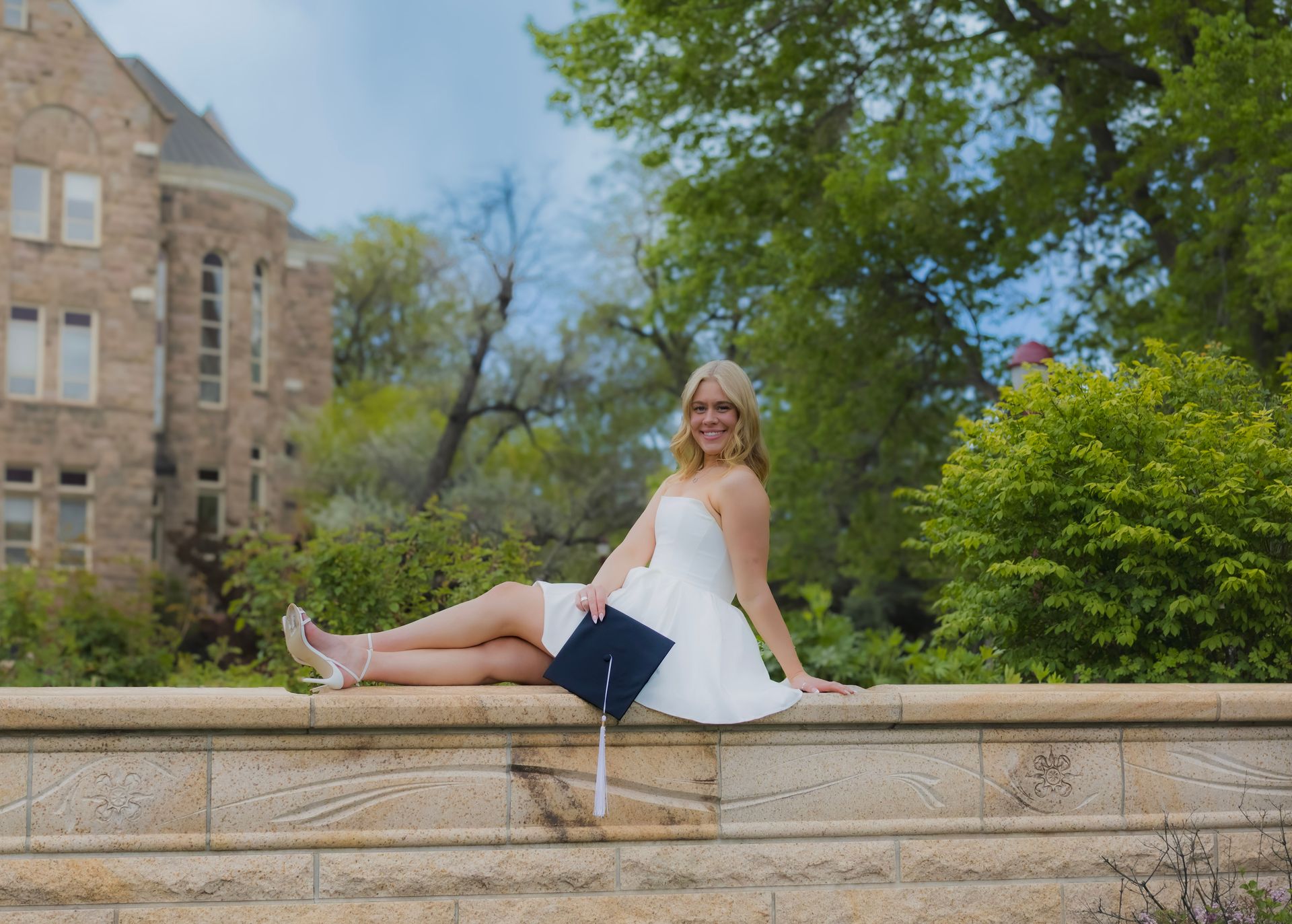 A woman in a white dress is sitting on a stone wall holding a graduation cap.