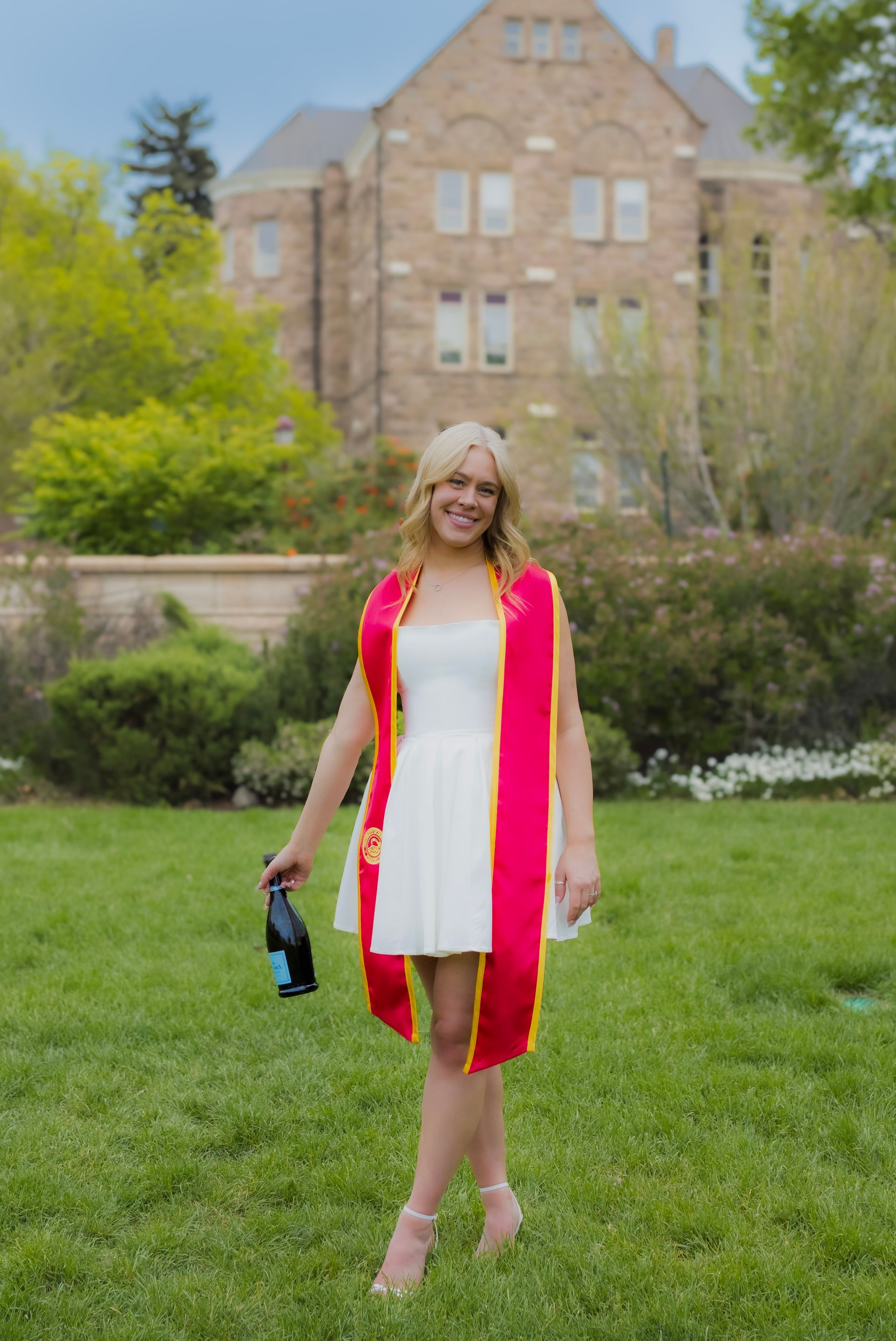 A woman in a red cape and white dress is standing in the grass in front of a building.