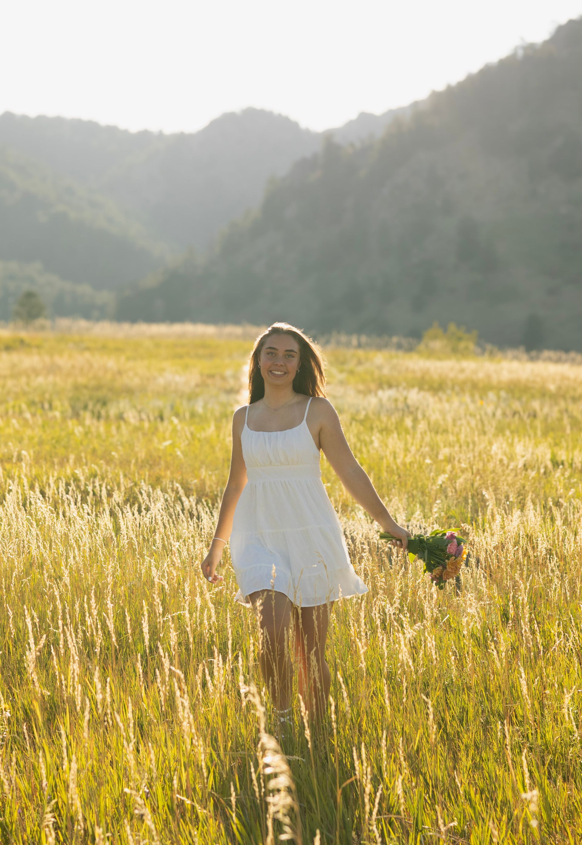 A woman in a white dress is walking through a field holding a bouquet of flowers.