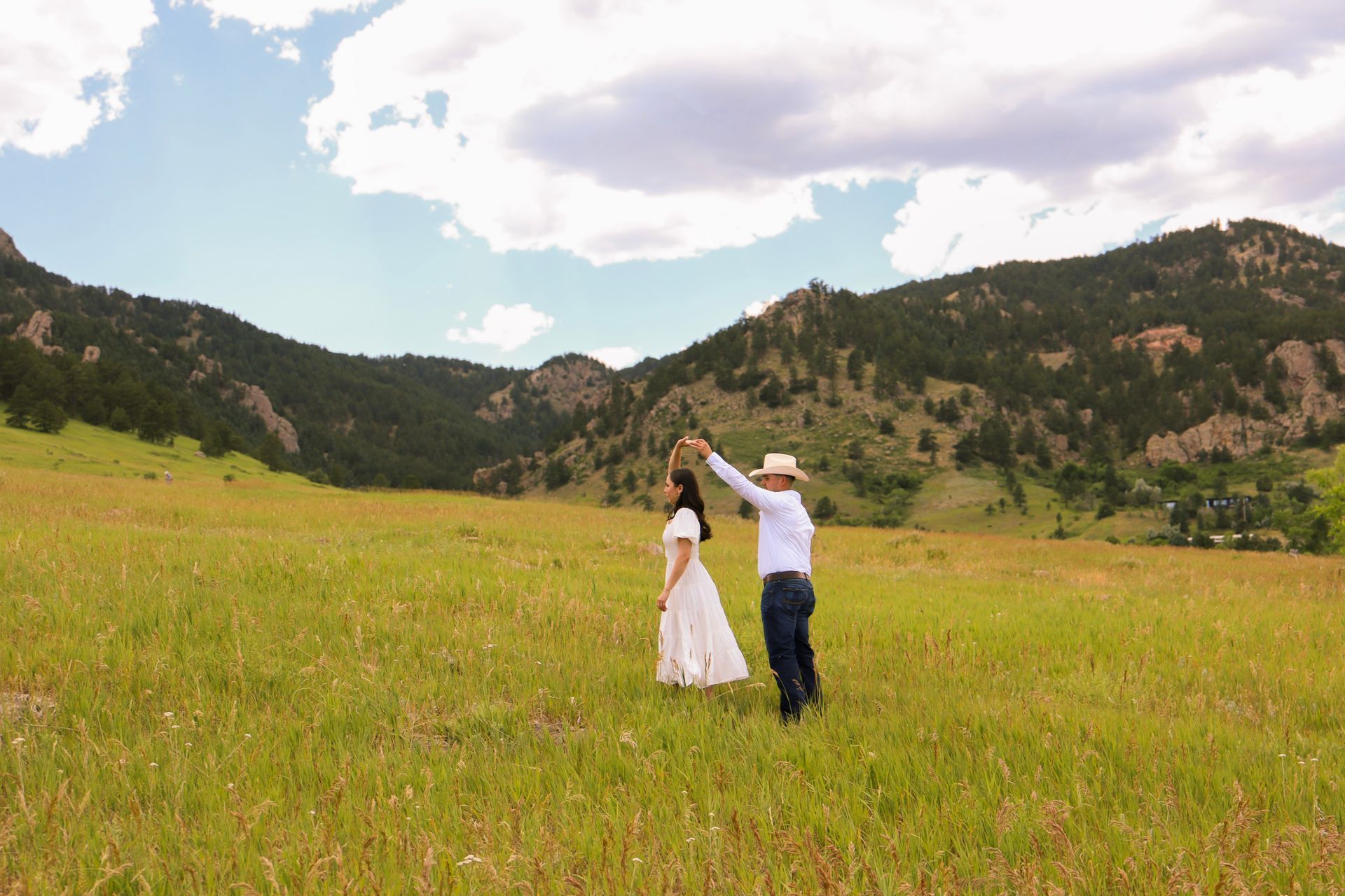 A man and a woman are dancing in a field with mountains in the background.