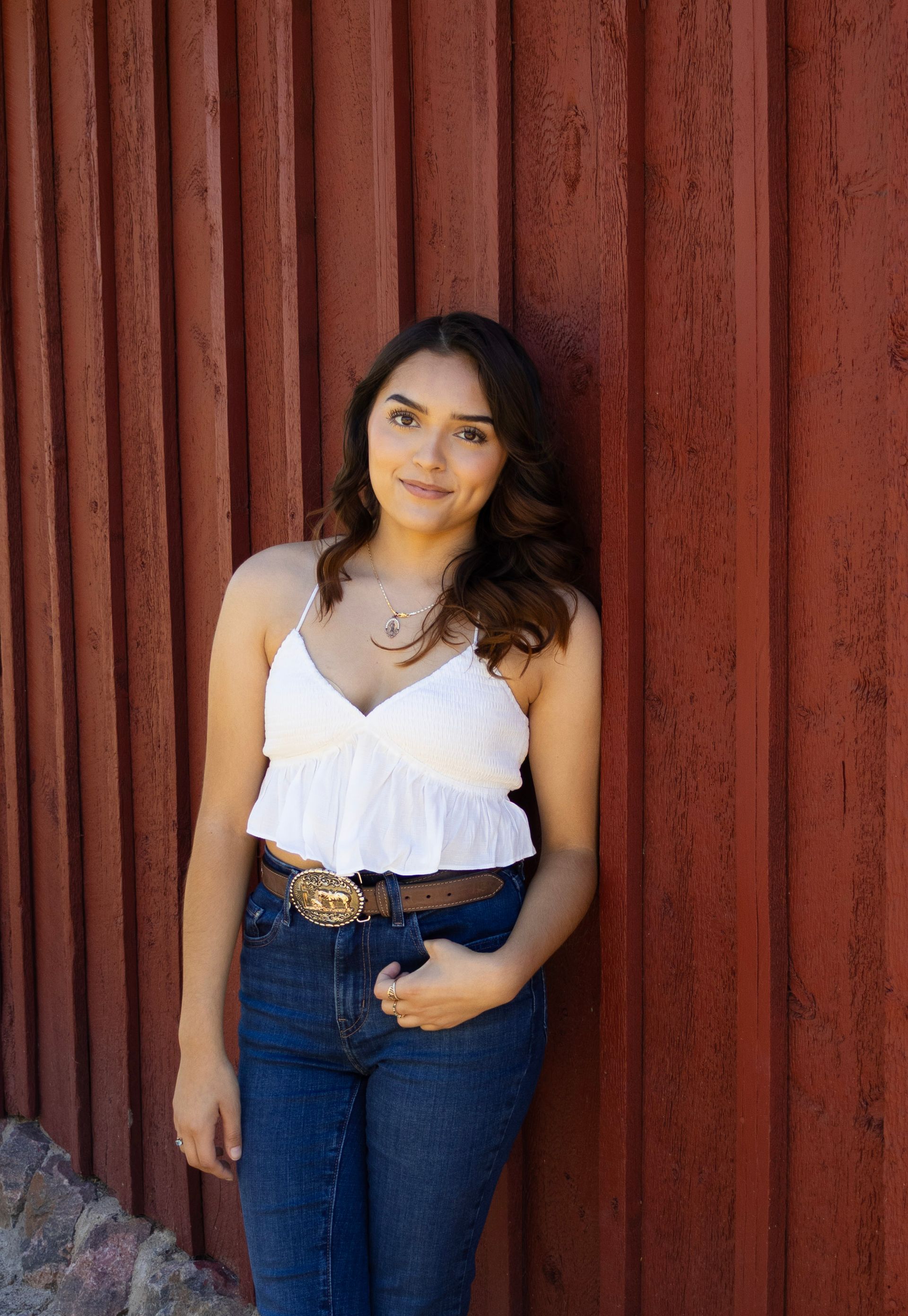 A woman in a white tank top and blue jeans is leaning against a red fence.