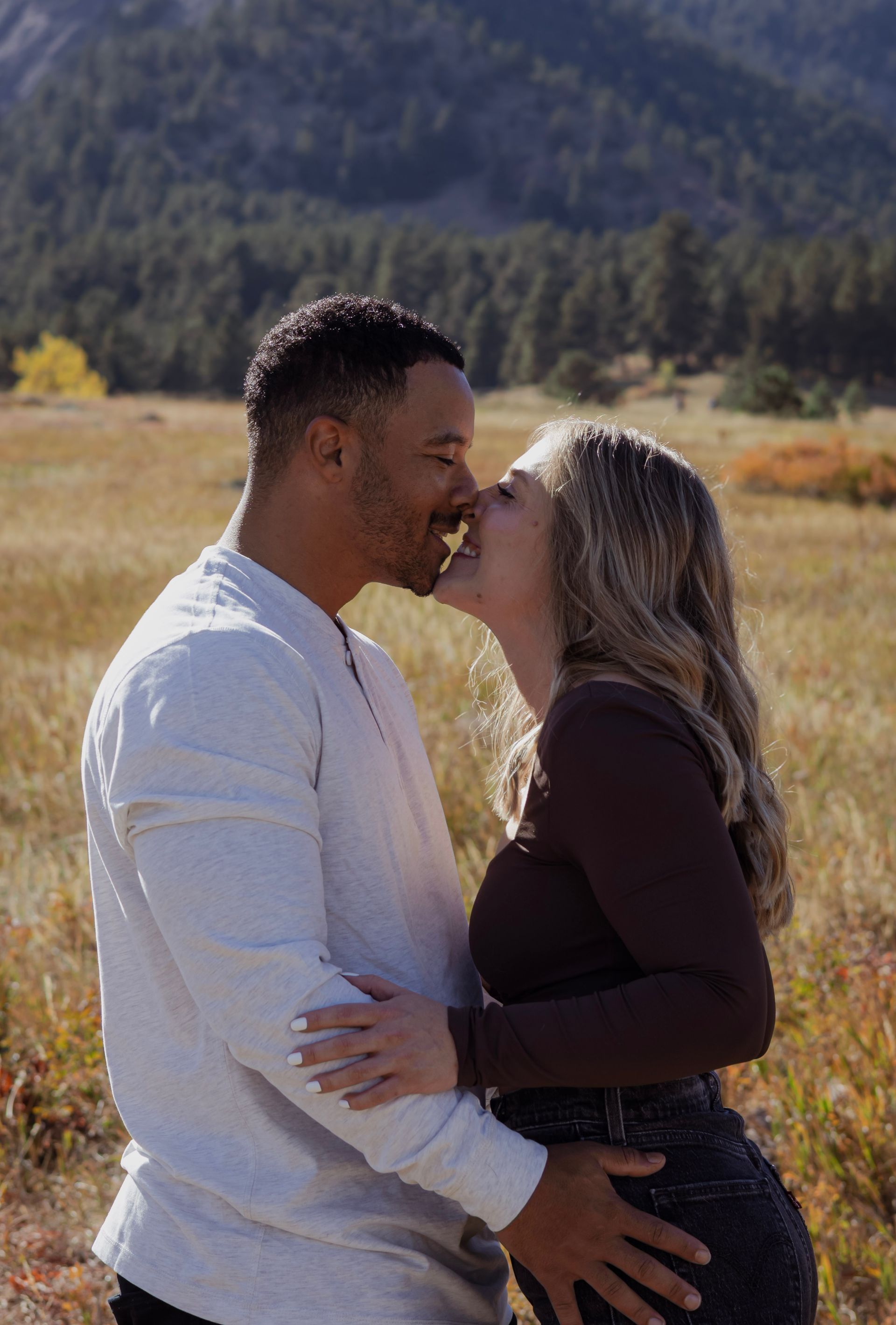 A man and a woman are kissing in a field with mountains in the background.