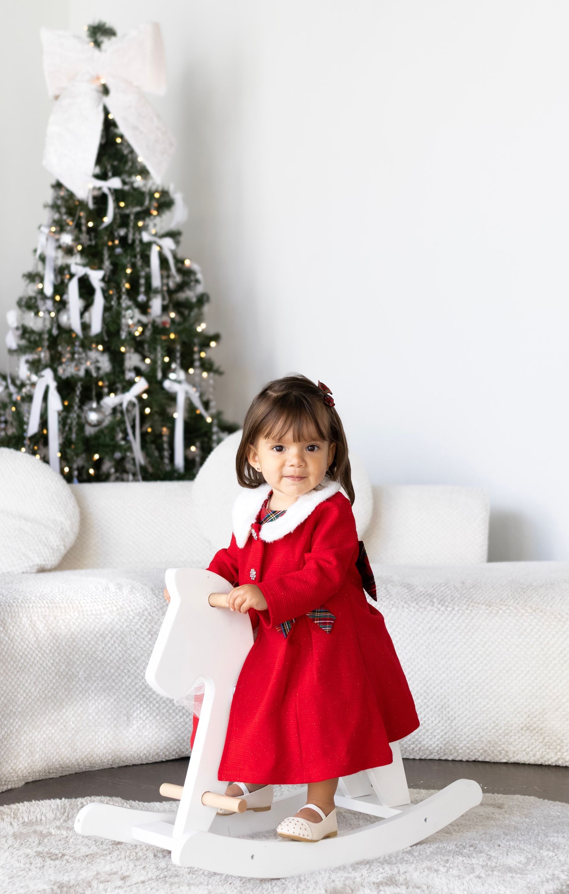 A little girl in a red dress is riding a rocking horse in front of a christmas tree.