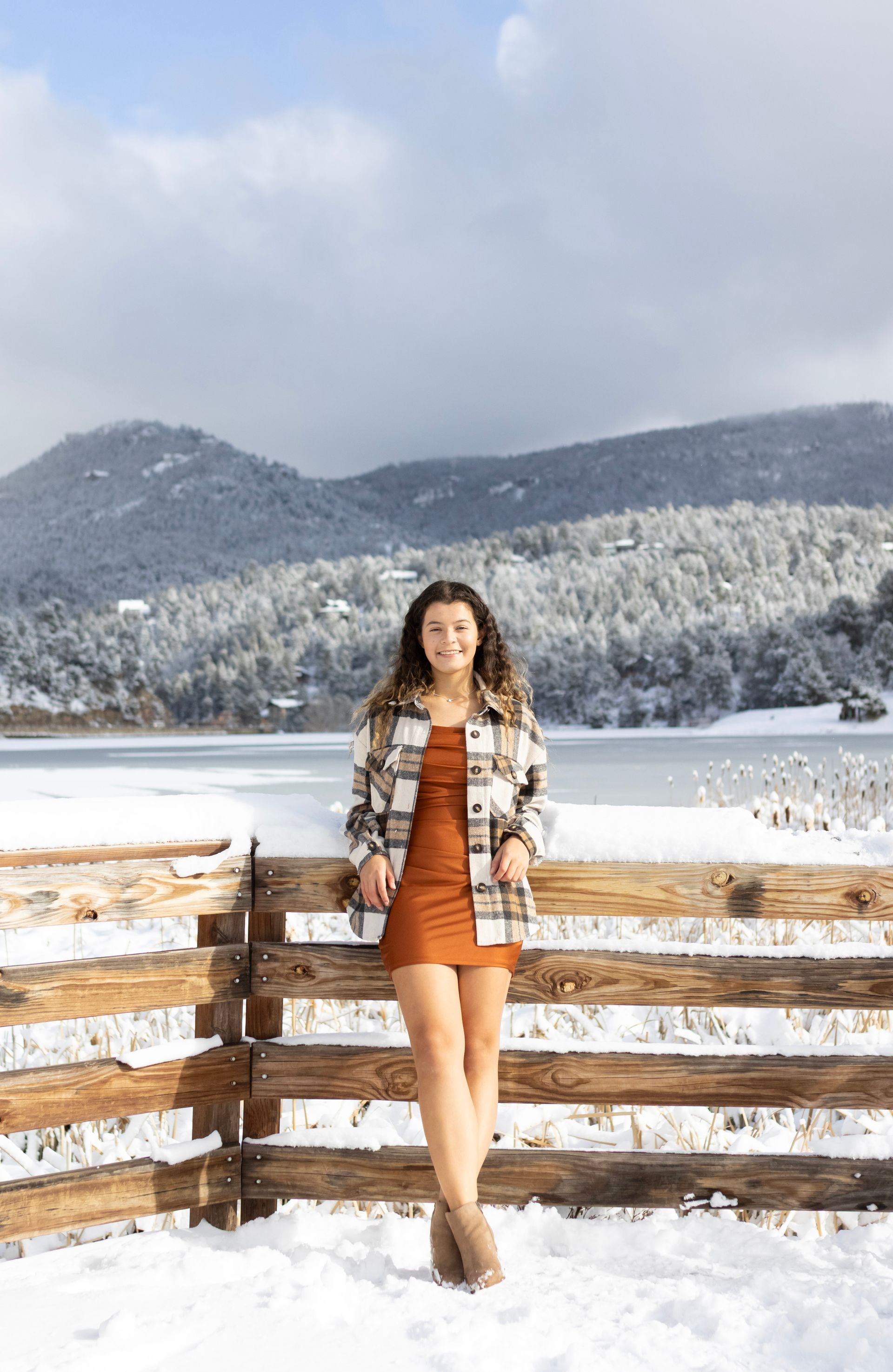 A woman in an orange dress is leaning against a wooden fence in the snow.