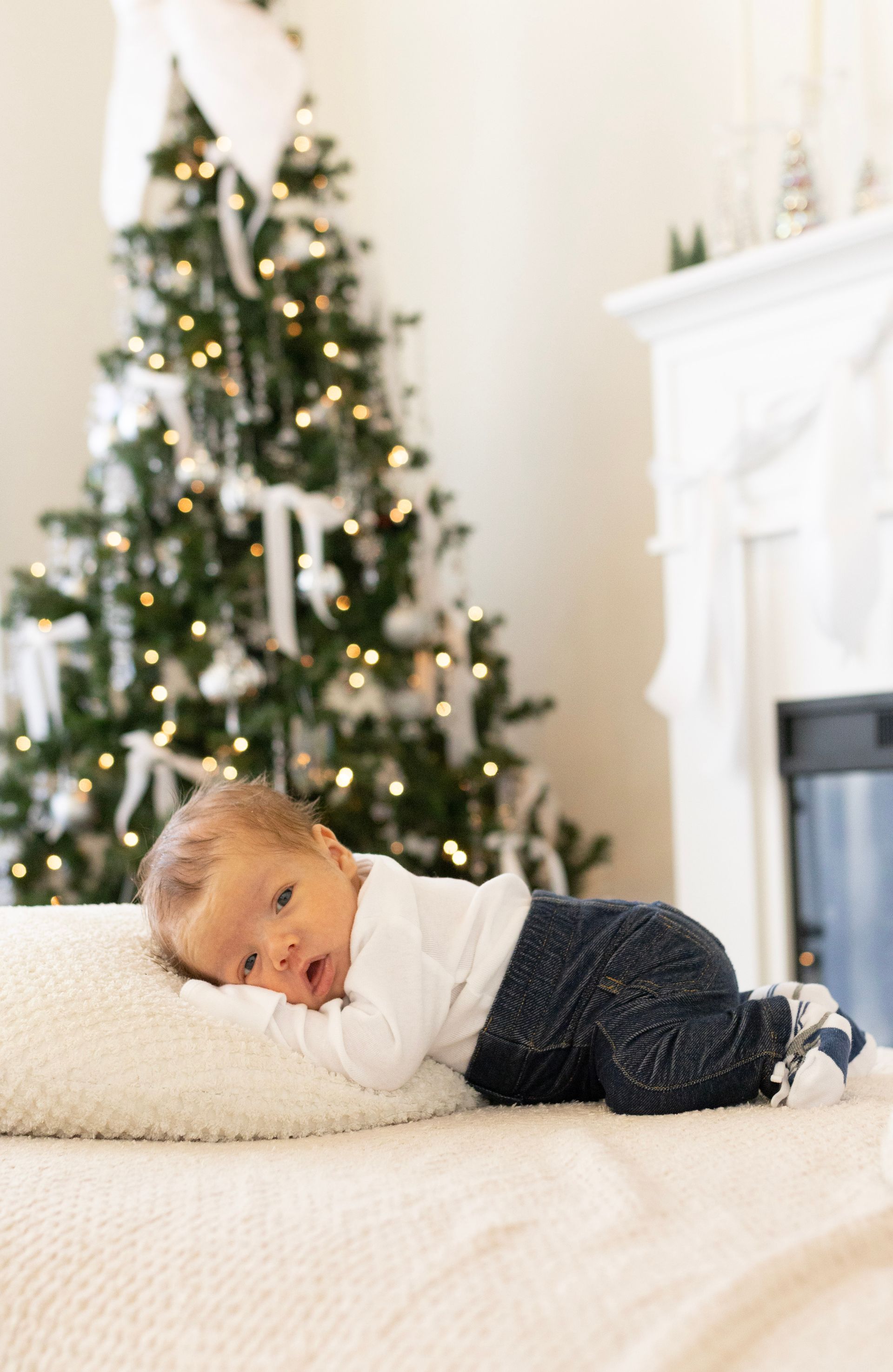 A baby is laying on a bed in front of a christmas tree.