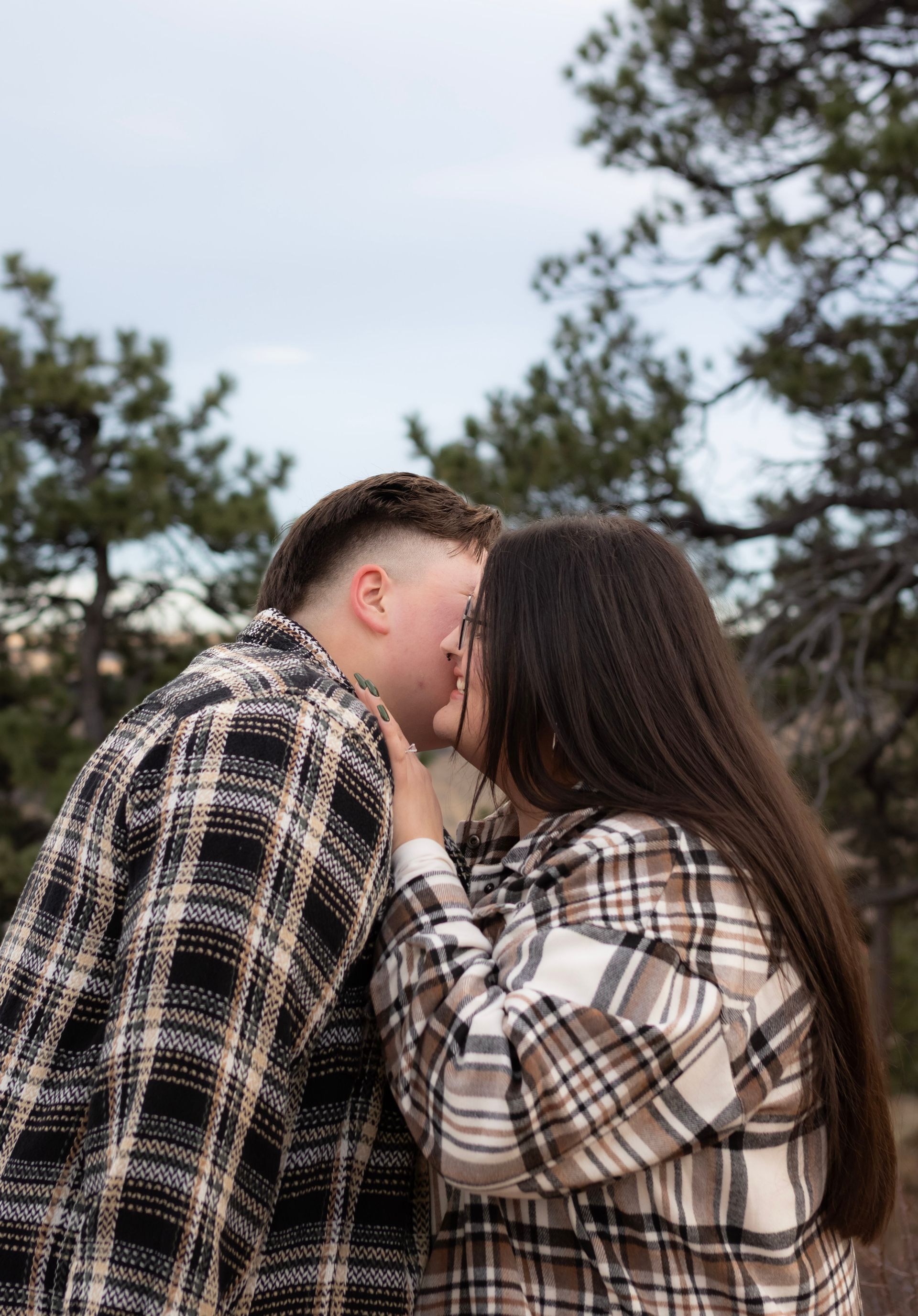 A man and a woman are kissing in front of trees.