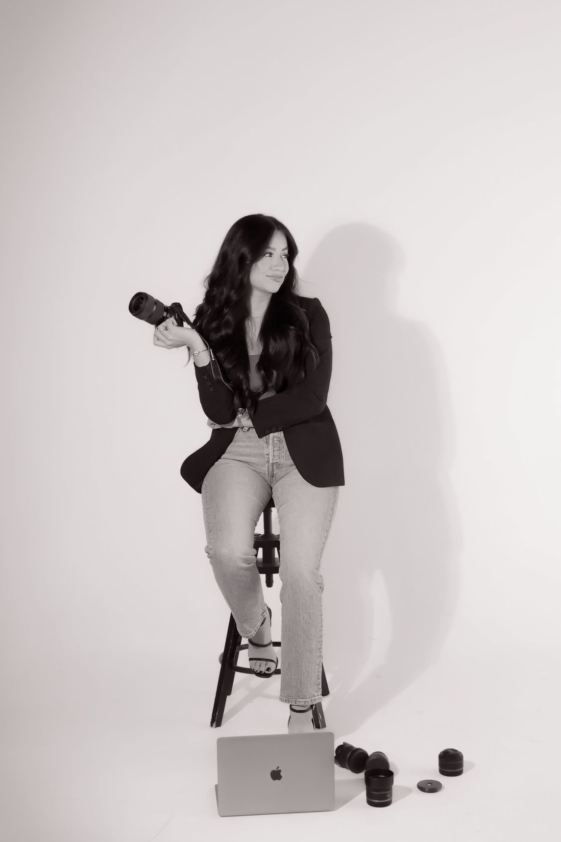 A woman is sitting on a stool holding a camera and a laptop.