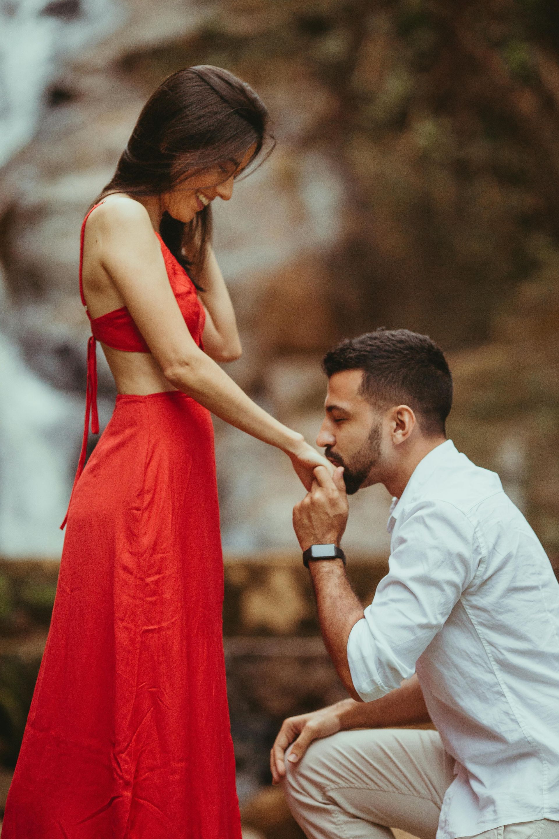 A man is kissing a woman 's hand in front of a waterfall.