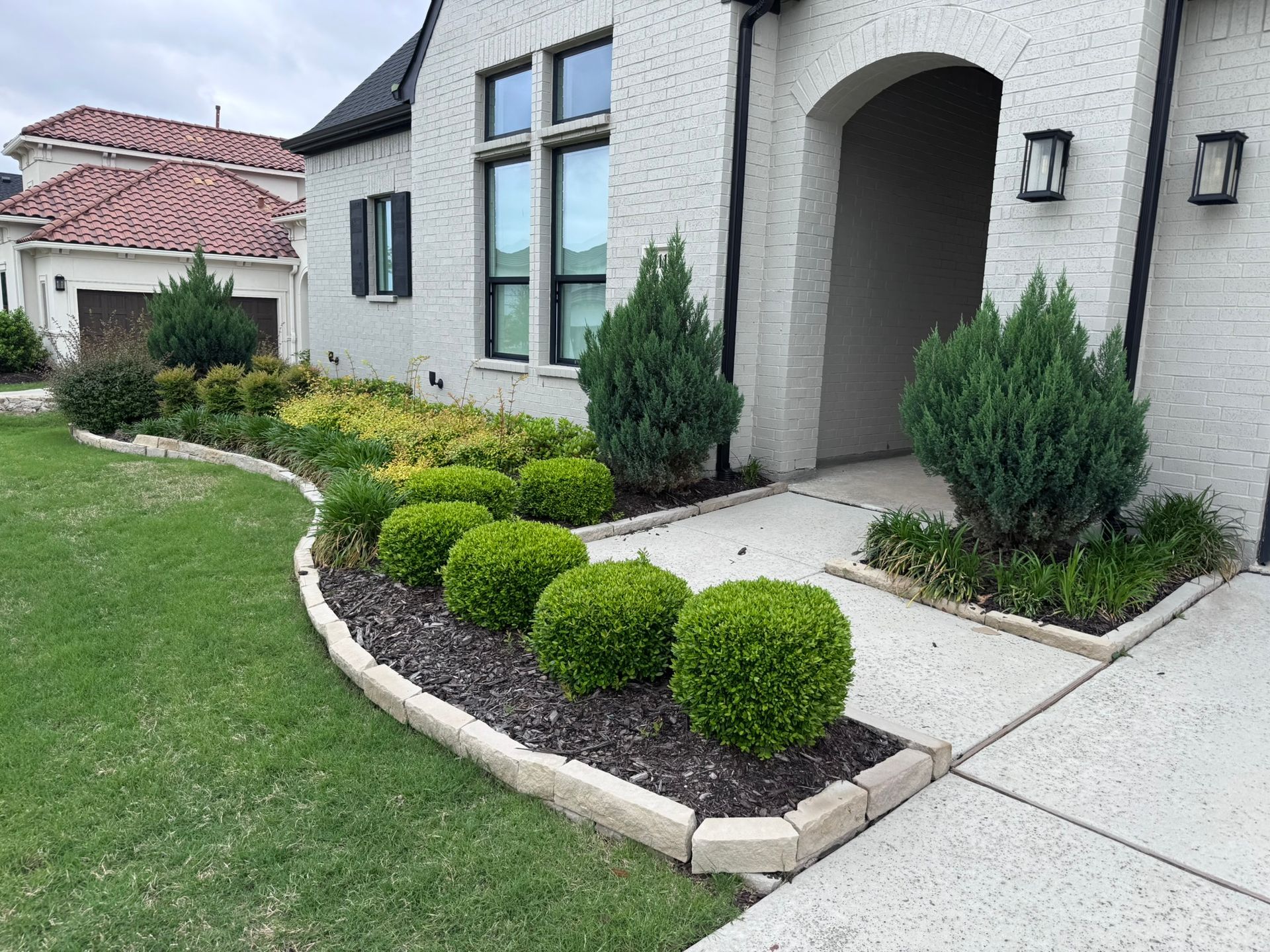 A well-landscaped front yard with a brick house and a curving flower bed filled with green shrubs and mulch.