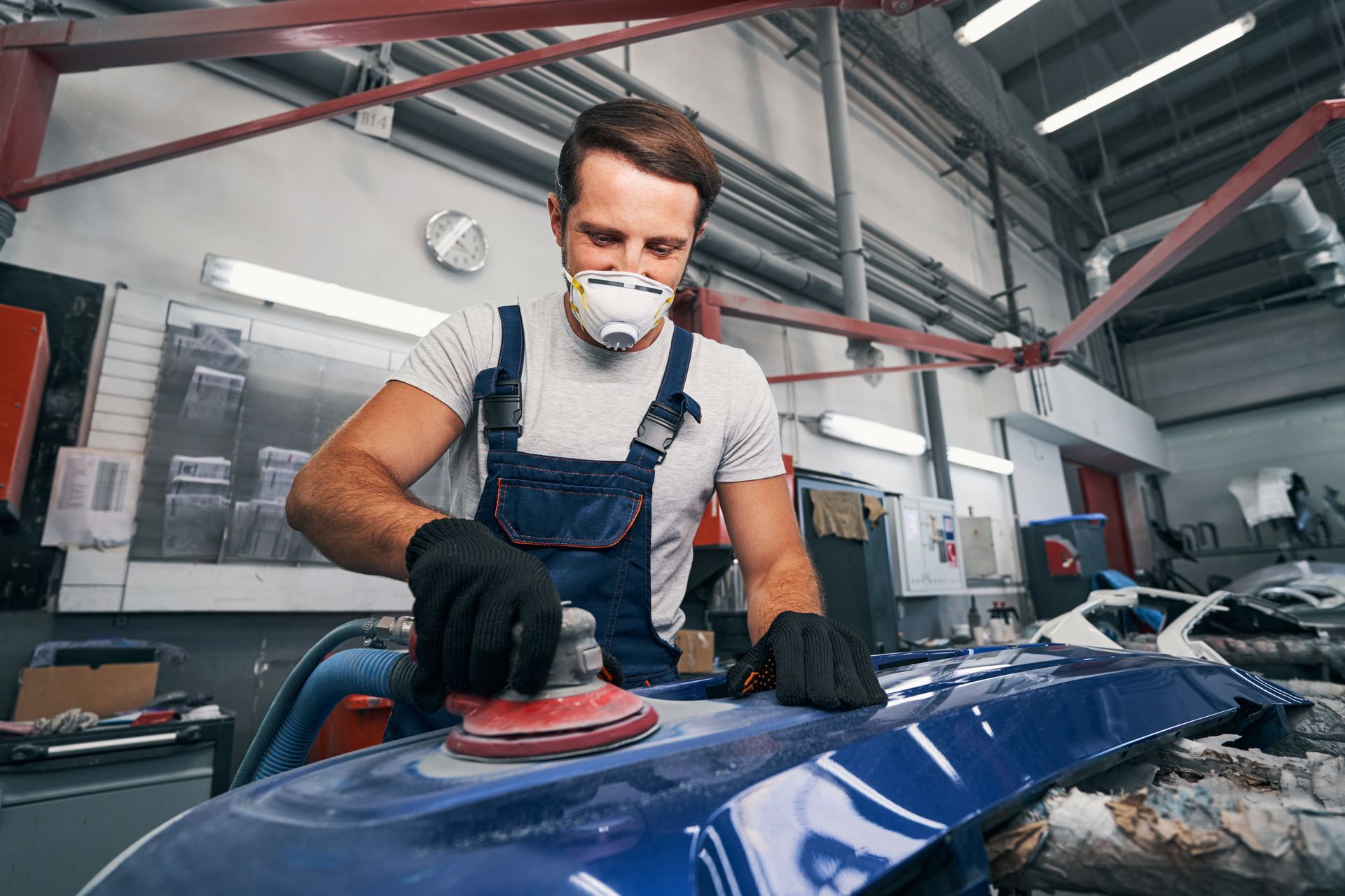 Auto body worker sanding a car panel with a power sander inside a repair shop.