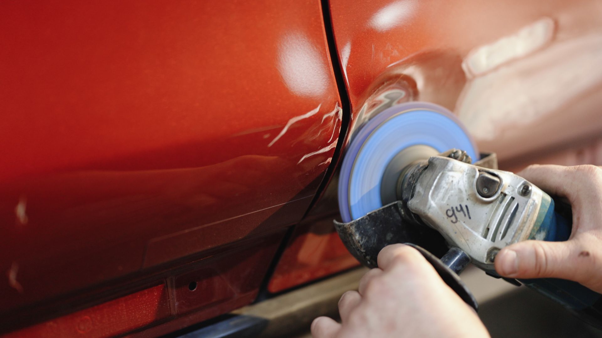 Hands using a power grinder to smooth damage on a red car panel in an auto body shop.