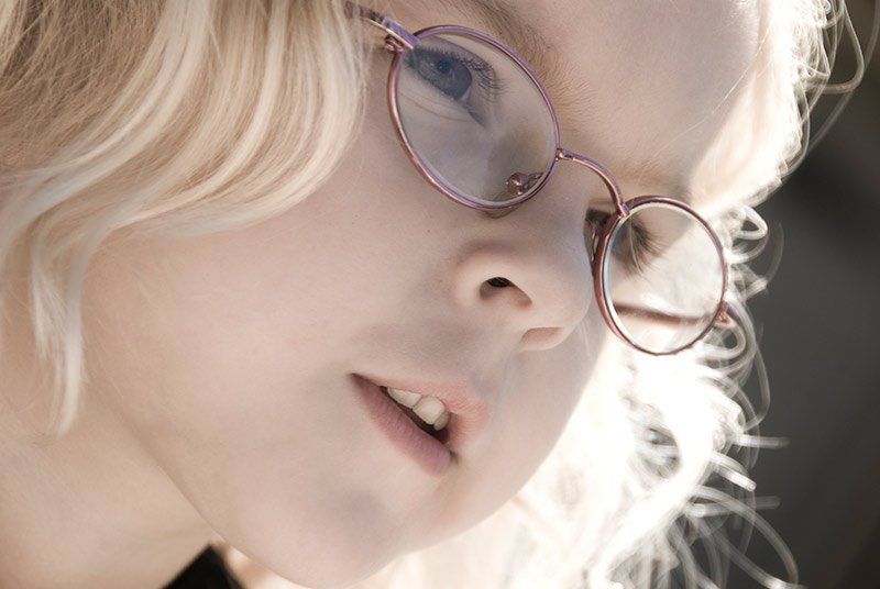 Blond child with glasses looking down, light reflecting on face.
