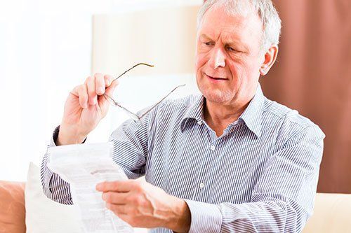 Older man holding glasses and document, squinting with concerned expression.