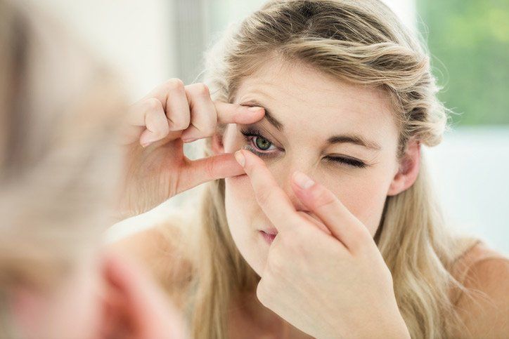 Woman in front of a mirror, inserting contact lens in her eye. She is winking.