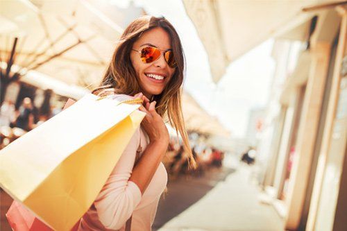 Woman smiling, holding shopping bags, walking down a sunny street, wearing sunglasses.