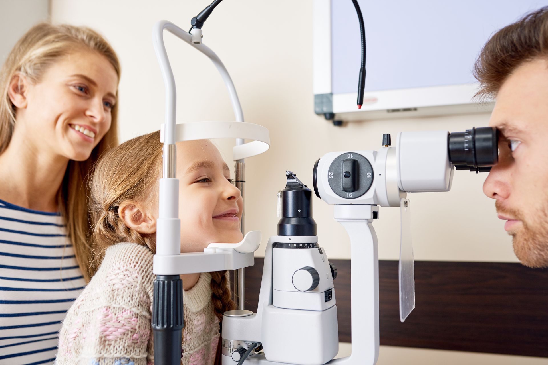 A child is receiving an eye exam using a slit lamp while an adult stands nearby.