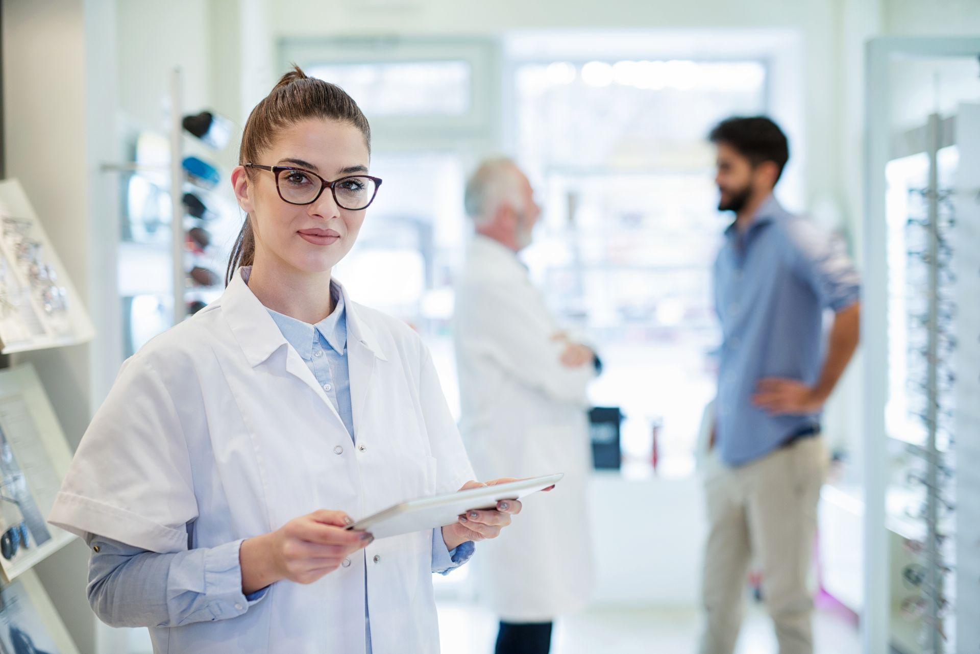 Optician holding a tablet in an eyewear shop with staff and a customer in the background. 
