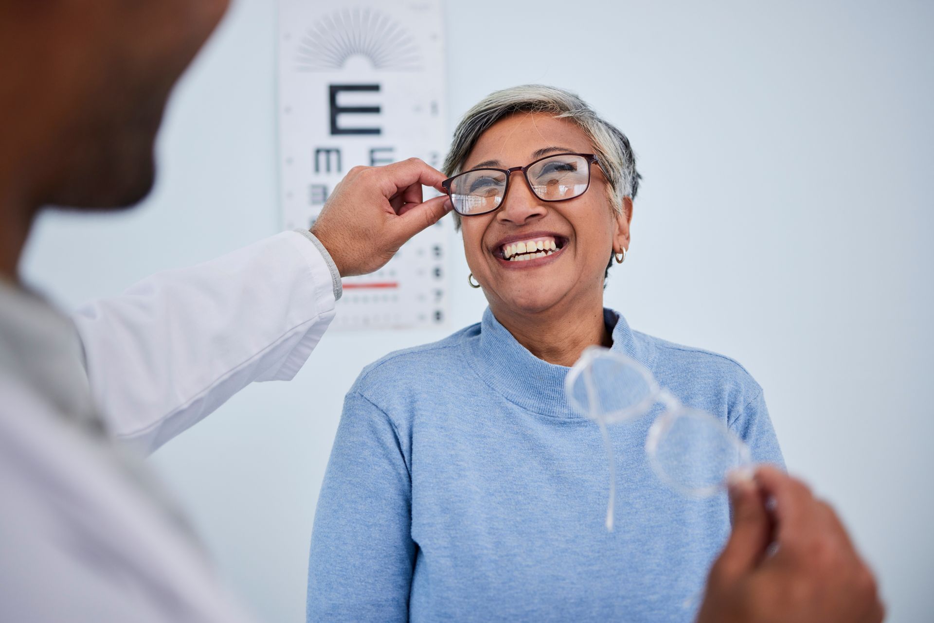 Healthcare professional fitting eyeglasses on a patient in an exam room.