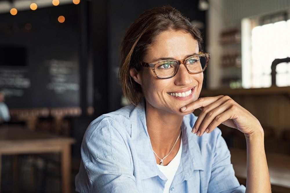 Woman with glasses smiles, resting chin on hand in a café setting.