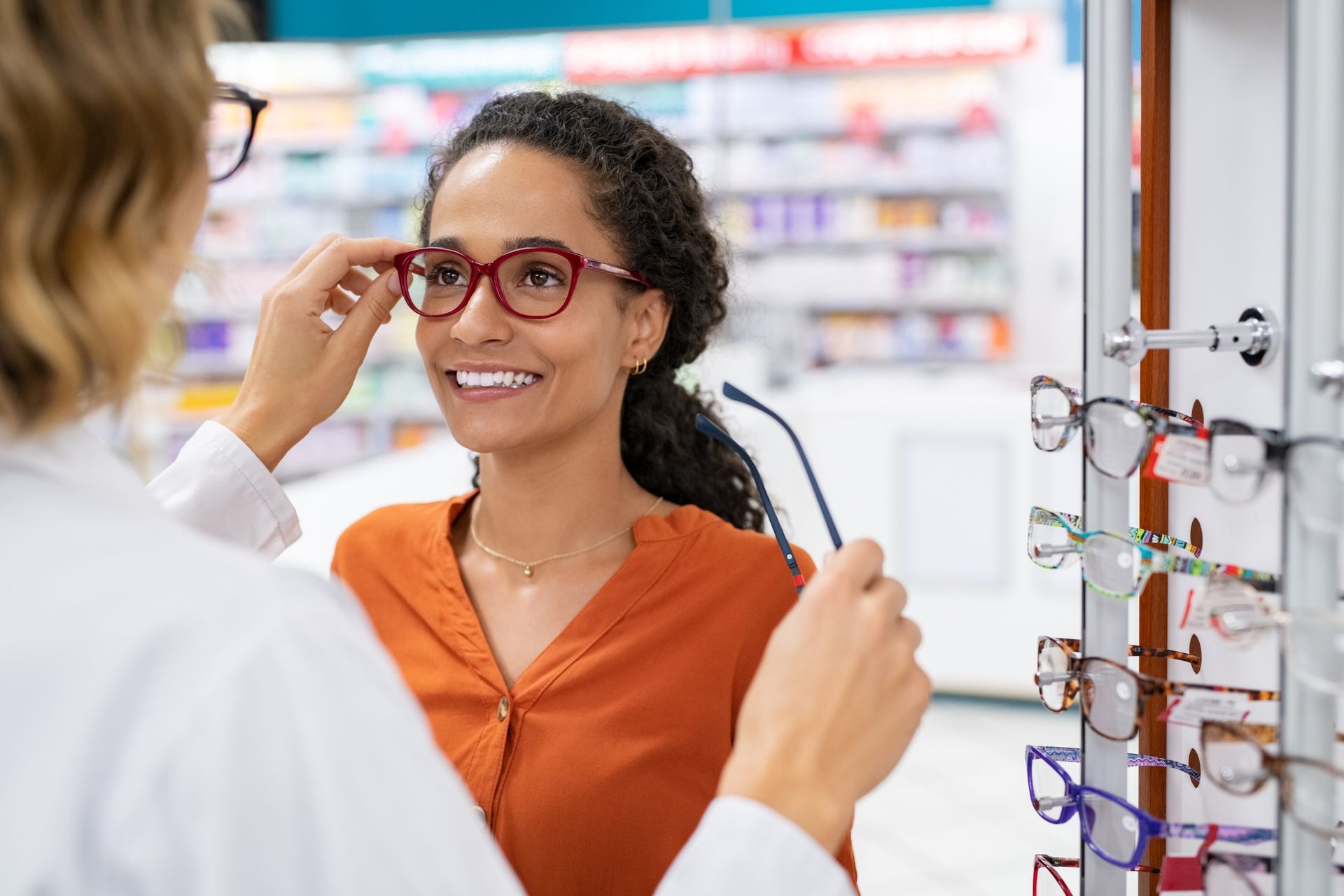 Optician helping a customer try on red eyeglasses in a store. Customer smiles.