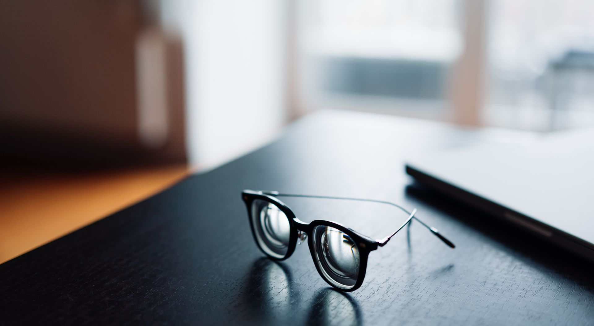 Eyeglasses on a dark table, near a blurred laptop and window. Eyeglasses on a dark table, near a blurred laptop and window.