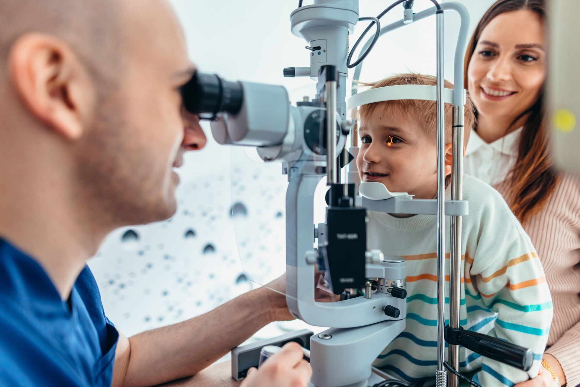 Optometrist examining a child's eyes with a slit lamp; the mother watches.