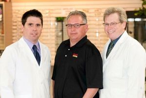 Three men, two in white coats, one in black shirt, stand in a shop with glasses displayed.