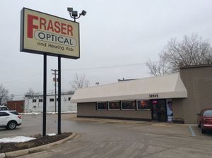 Fraser Optical and Hearing Aids store with sign. Brown building, black poles, cloudy sky, cars in parking lot.