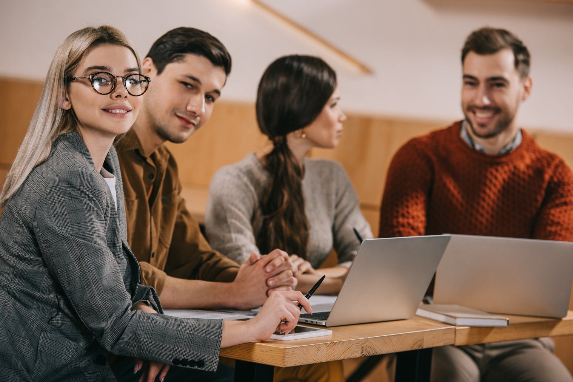 Four people at a table with laptops, one woman in glasses smiles, others look at her, all in a cafe setting. Four people at a table with laptops, one woman in glasses smiles, others look at her, all in a cafe setting.