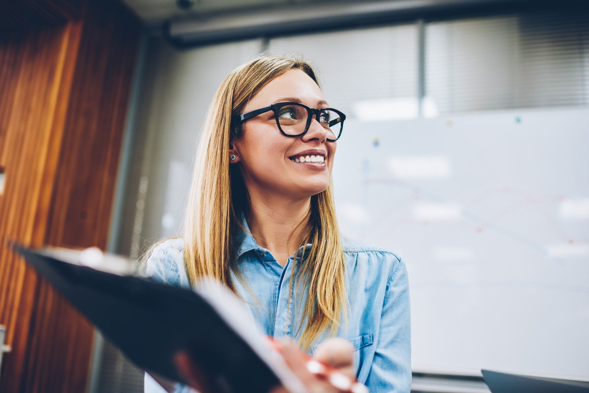 Woman wearing glasses, smiling, holding a binder, in front of a whiteboard. Woman wearing glasses, smiling, holding a binder, in front of a whiteboard.