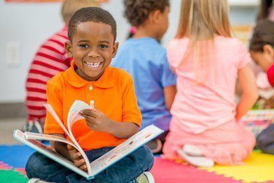 Boy smiles while reading a book in a classroom; children sit nearby. Boy smiles while reading a book in a classroom; children sit nearby.
