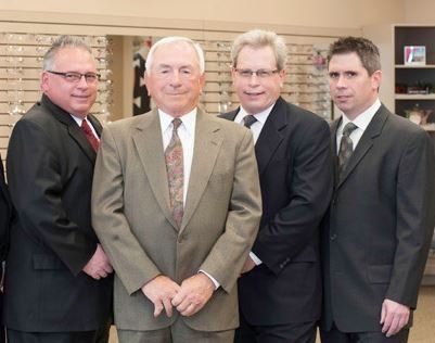 Four men in suits stand inside an eyewear store, smiling.
