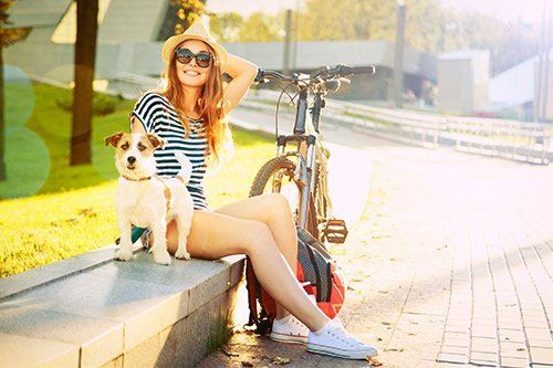 Woman sitting with dog beside bicycle, wearing hat and sunglasses, sunny outdoor setting.