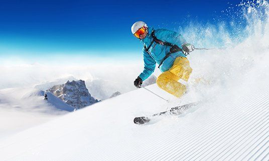 Skier in blue jacket, yellow pants, skis down a snowy slope, white powder spray, blue sky.