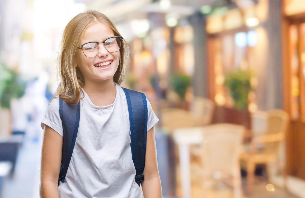 Girl with glasses and backpack smiles in front of a blurred outdoor cafe.