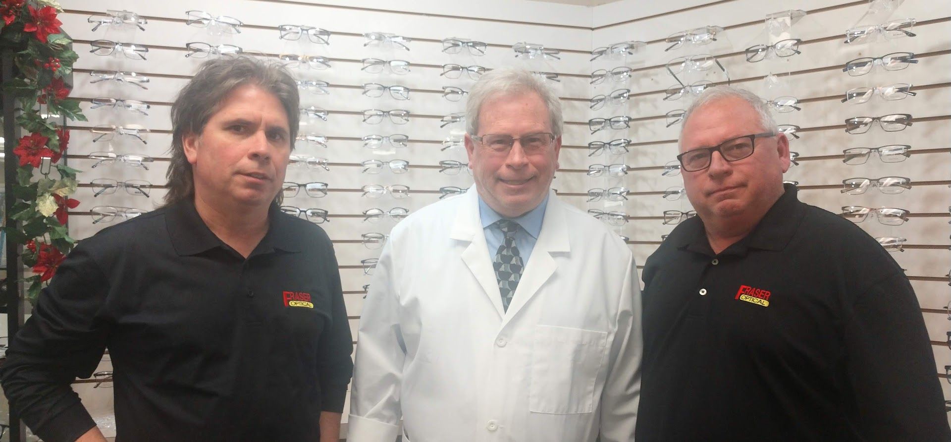 Three men standing in front of a wall of eyeglass frames, one in a white coat and two in black shirts