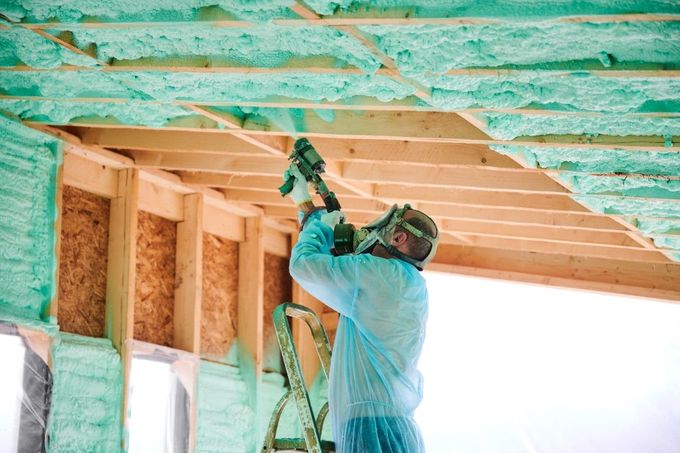 Construction worker spraying green foam insulation into a wooden ceiling, wearing protective gear.