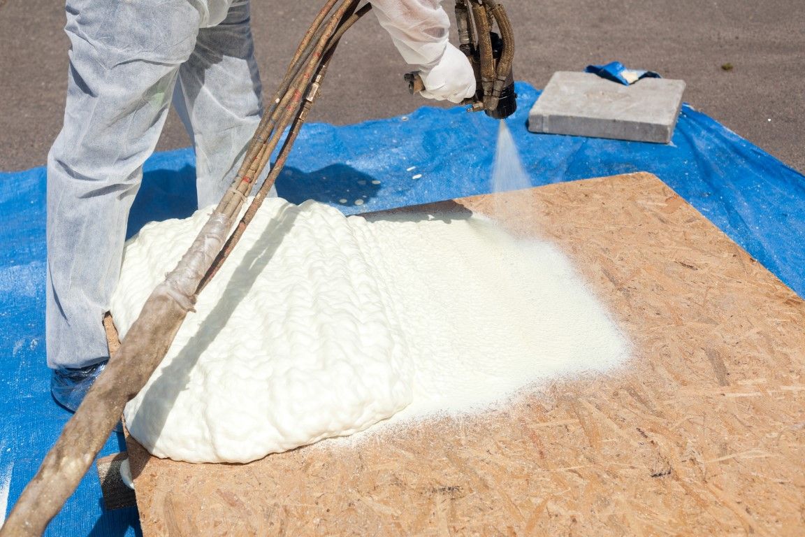 Person spraying white foam insulation onto a wooden board on a blue tarp.