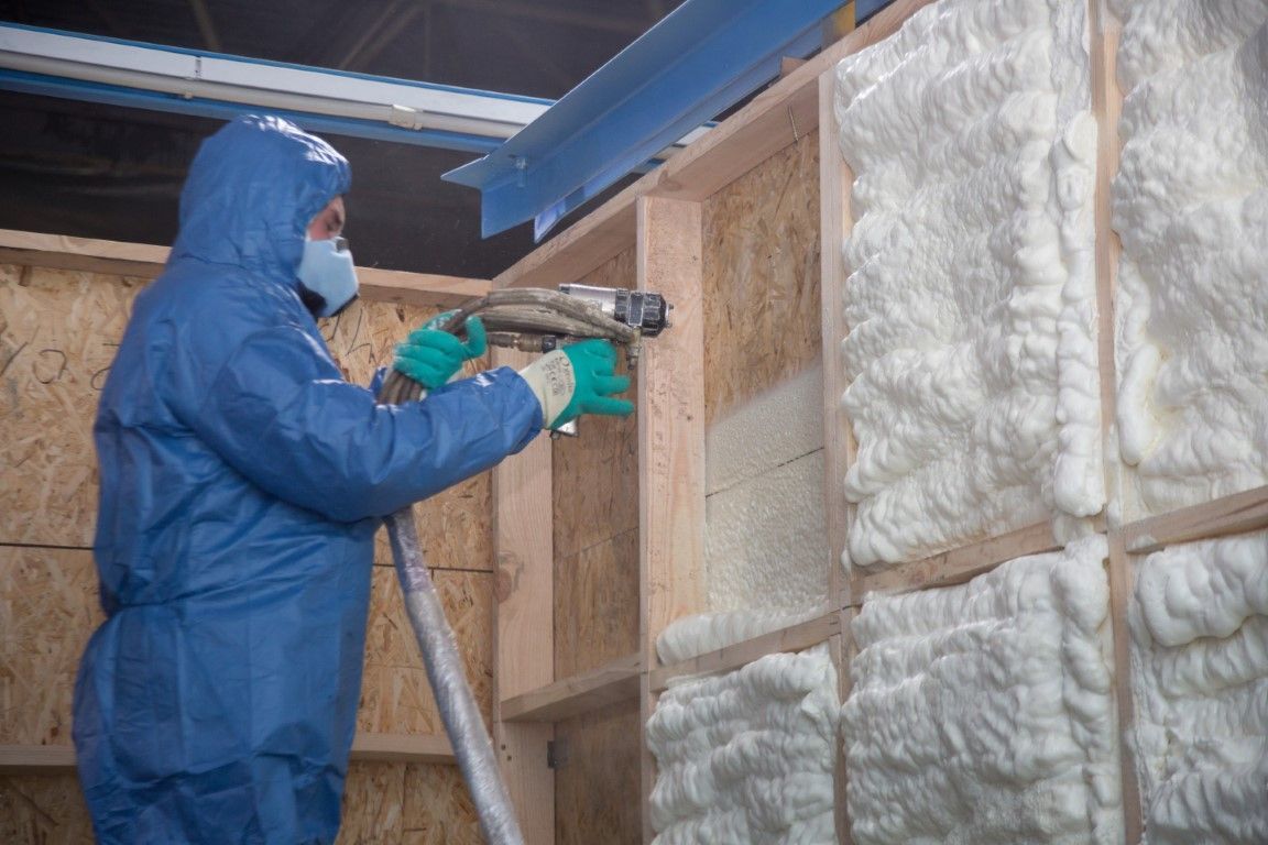 Person in protective suit spraying insulation foam onto a wall in a construction setting.