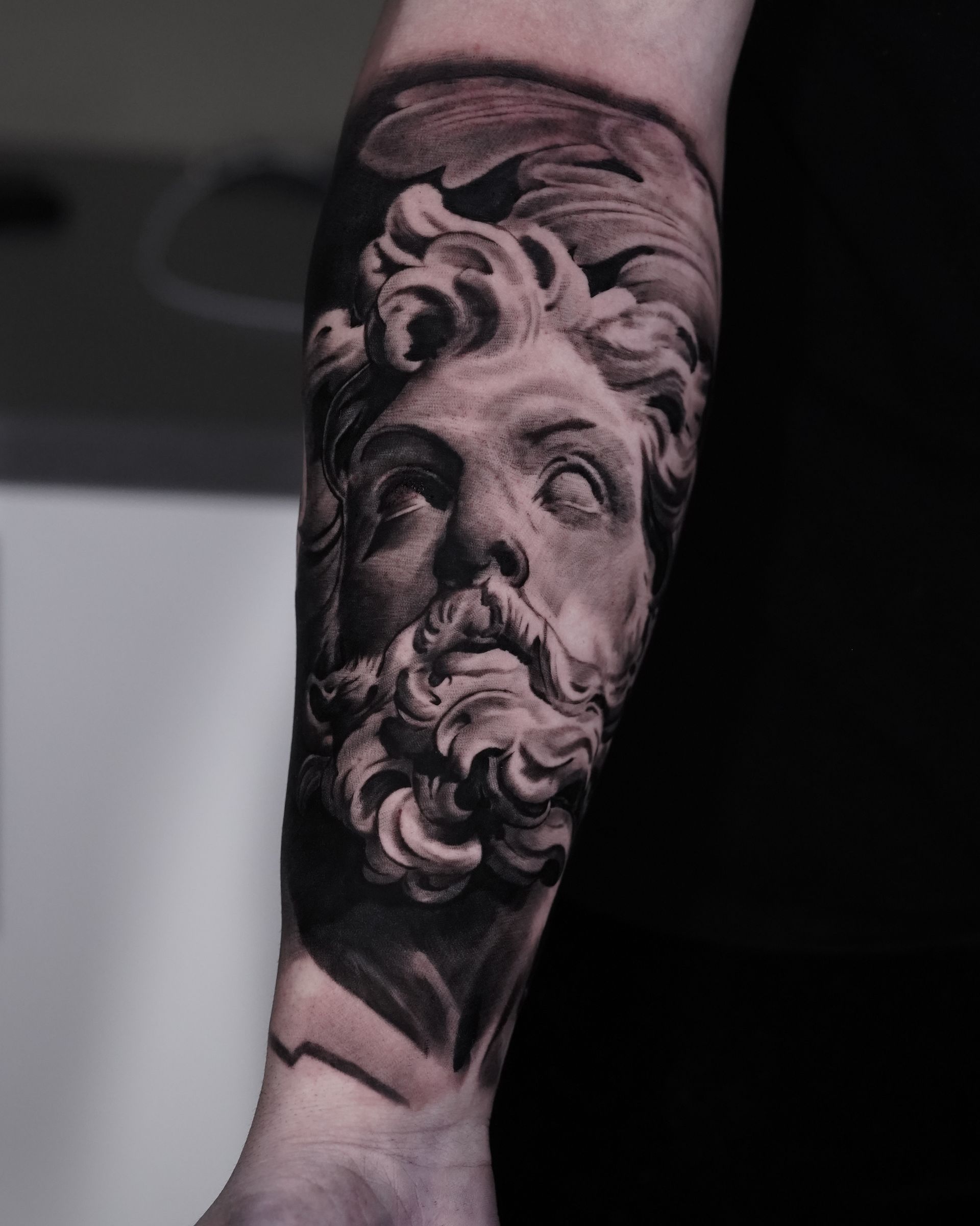 A man has a tattoo of a statue of a man with a beard on his forearm.